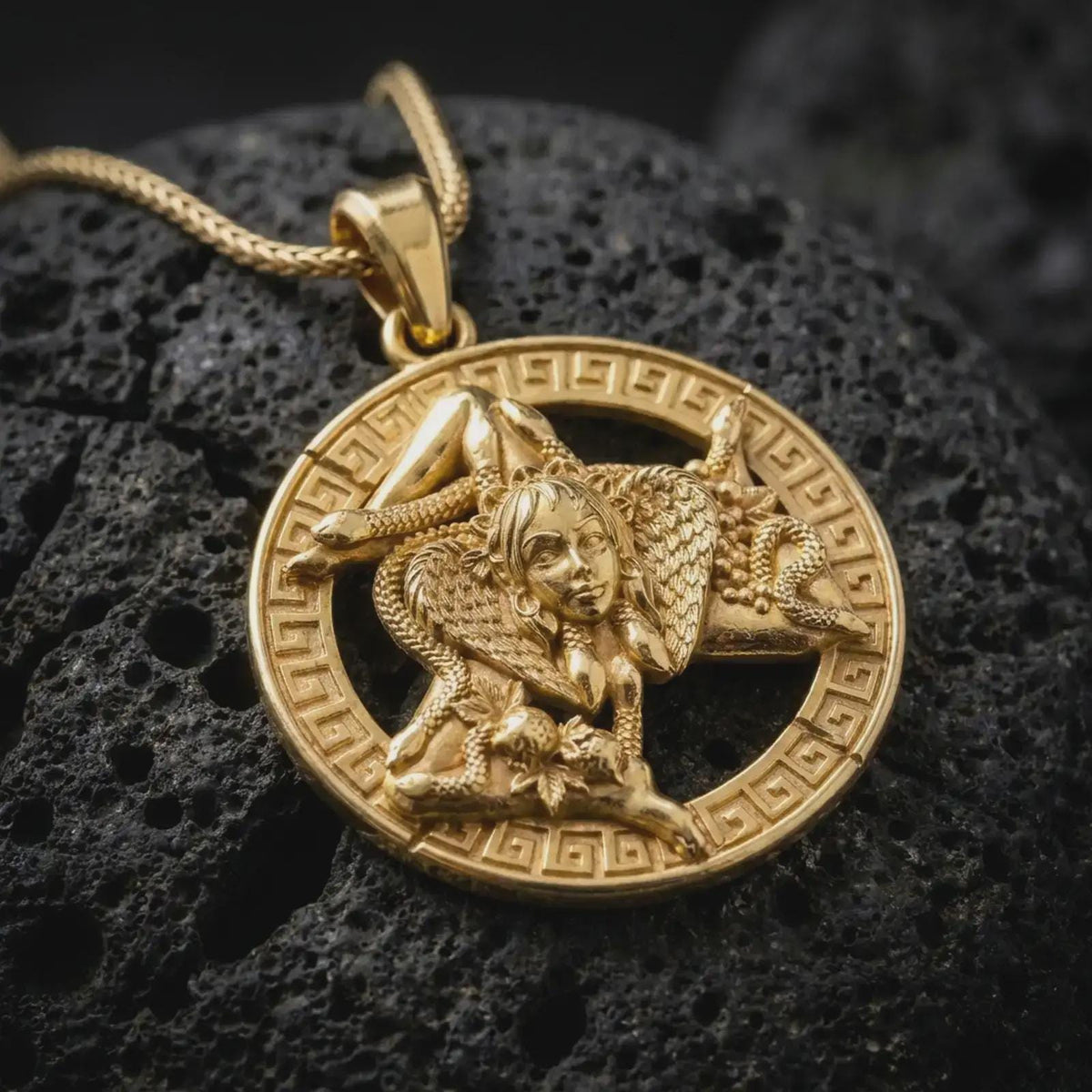 Gold plated Trinacria pendant displayed on dark volcanic rock, featuring the traditional Sicilian symbol with Medusa, three radiating legs, and an ornamental Greek key border.