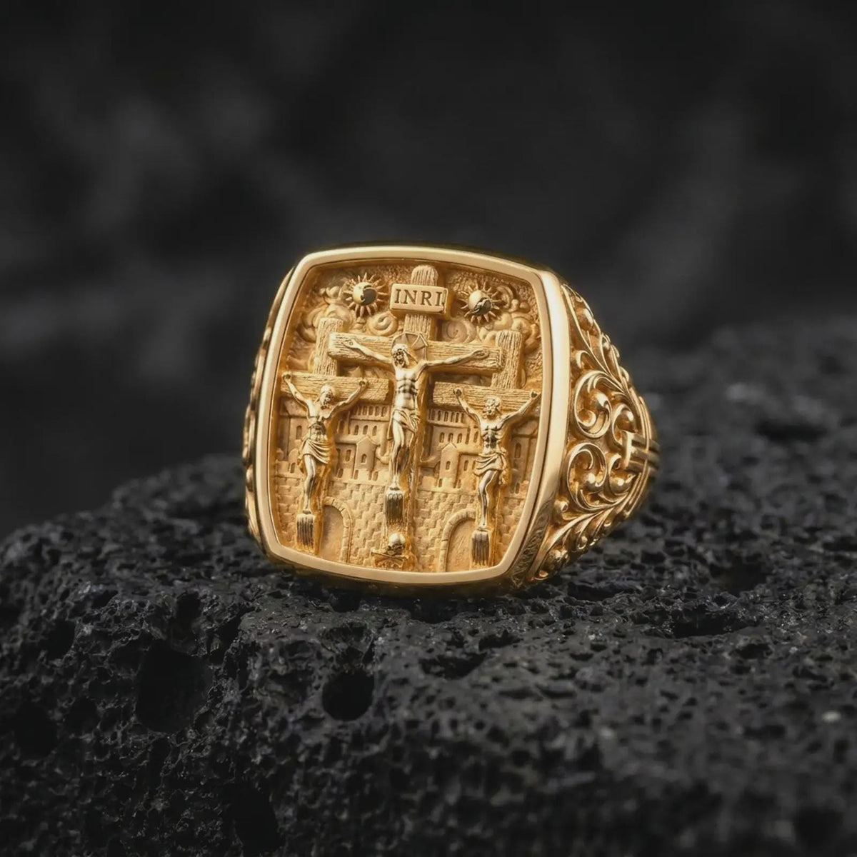 Gold plated Crucifixion ring resting on black volcanic rock, featuring Jesus Christ at Calvary between the two thieves, INRI inscription, three crosses, and detailed religious scenery.