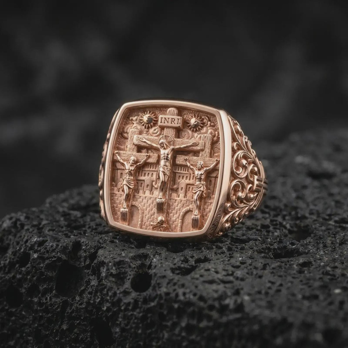 Rose gold plated Calvary ring displayed on textured volcanic stone, showcasing Jesus on the central cross, two crucified thieves, architectural details, and an ornate Christian signet profile.