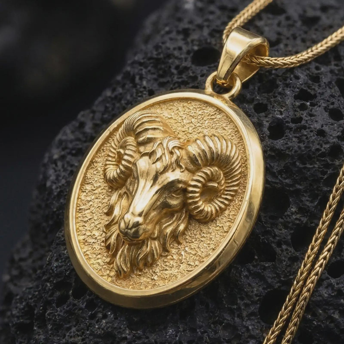 Gold plated Aries ram pendant resting on black volcanic rock, featuring a detailed ram head, powerful spiral horns, textured medallion background, and bold zodiac design.
