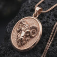Load image into Gallery viewer, Rose gold plated ram necklace displayed on textured volcanic stone, showcasing the sculpted Aries ram head, curled horns, facial details, and oval pendant profile.
