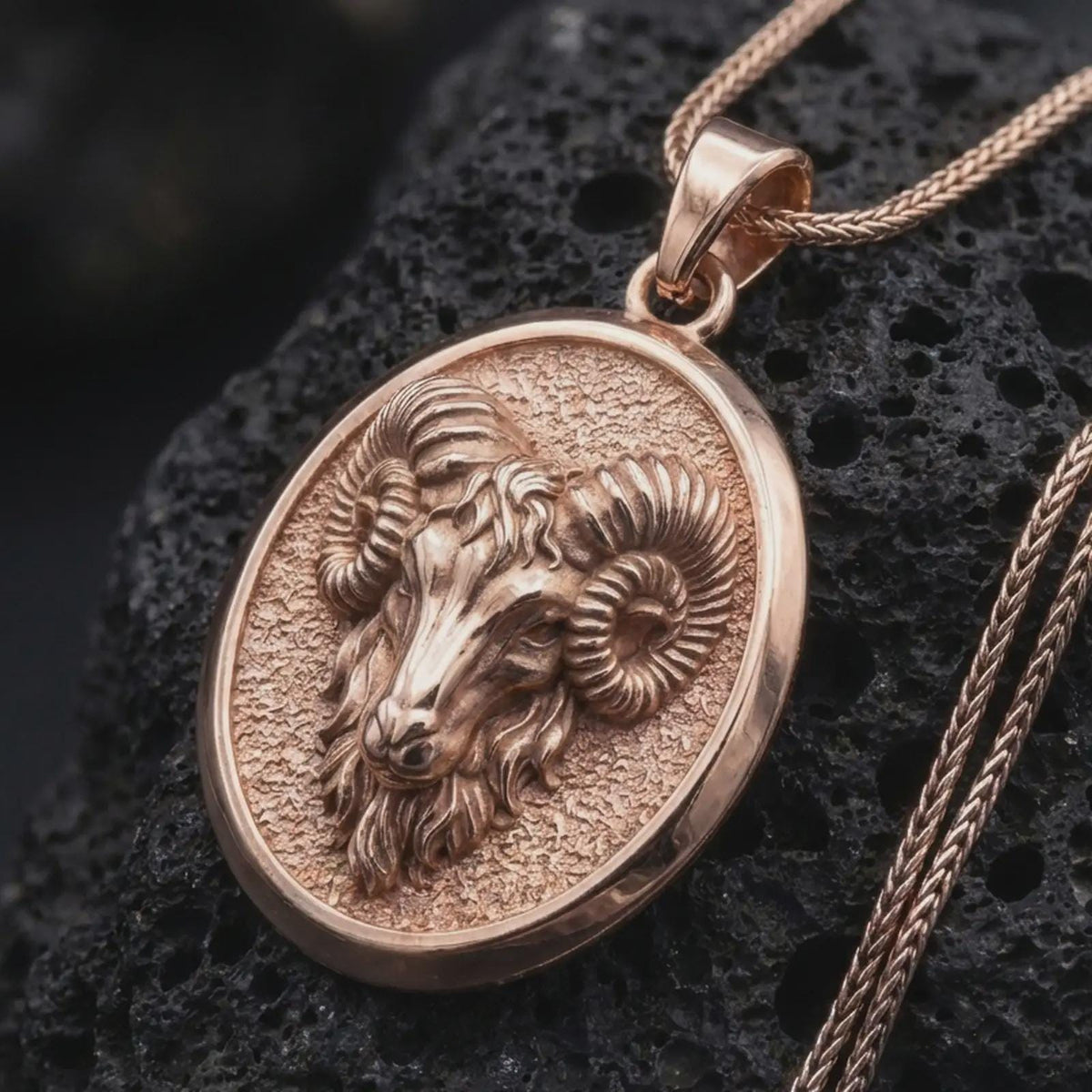 Rose gold plated ram necklace displayed on textured volcanic stone, showcasing the sculpted Aries ram head, curled horns, facial details, and oval pendant profile.