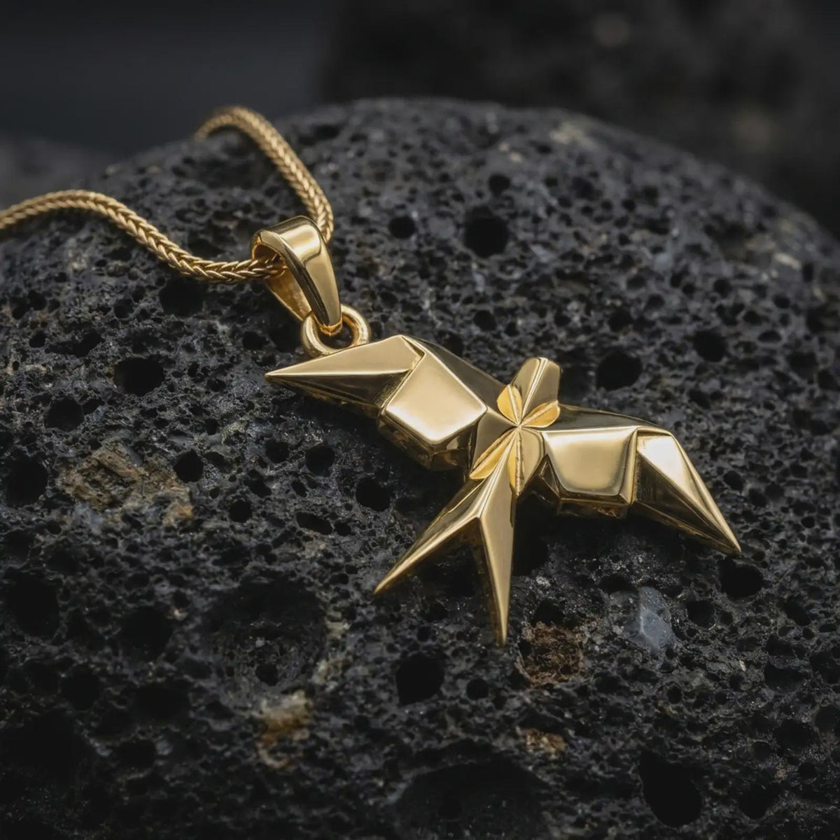 Gold plated origami swallow pendant resting on black volcanic rock, featuring a stylized flying swallow silhouette, sharp wing points, and clean faceted details.