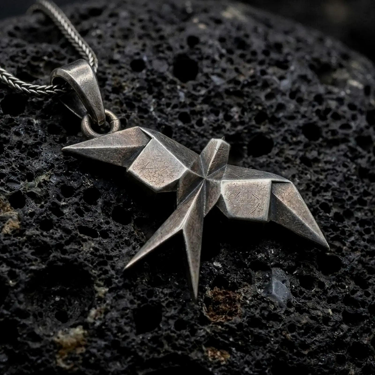 Oxidized sterling silver origami swallow pendant on black volcanic rock, featuring sharp geometric folds, pointed wings, and a faceted flying bird silhouette.