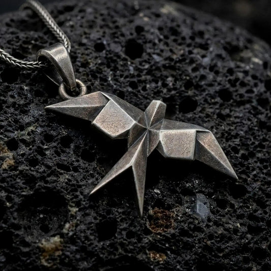 Oxidized sterling silver origami swallow pendant on black volcanic rock, featuring sharp geometric folds, pointed wings, and a faceted flying bird silhouette.