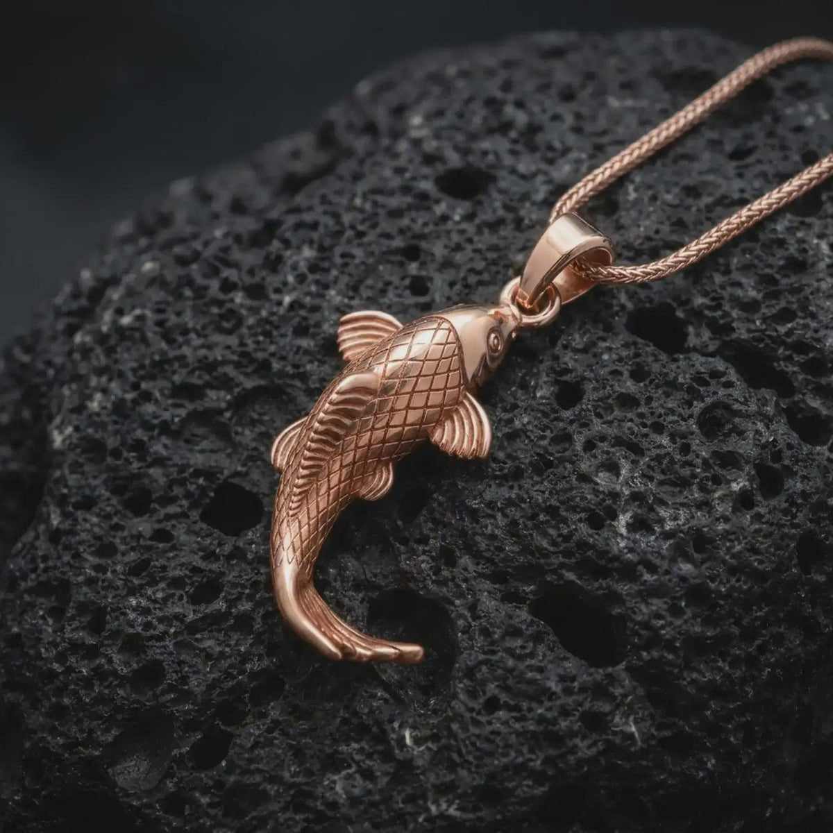 Rose gold plated koi necklace displayed on textured volcanic stone, showcasing the Japanese fish motif, engraved scales, flowing fins, and graceful dimensional body.