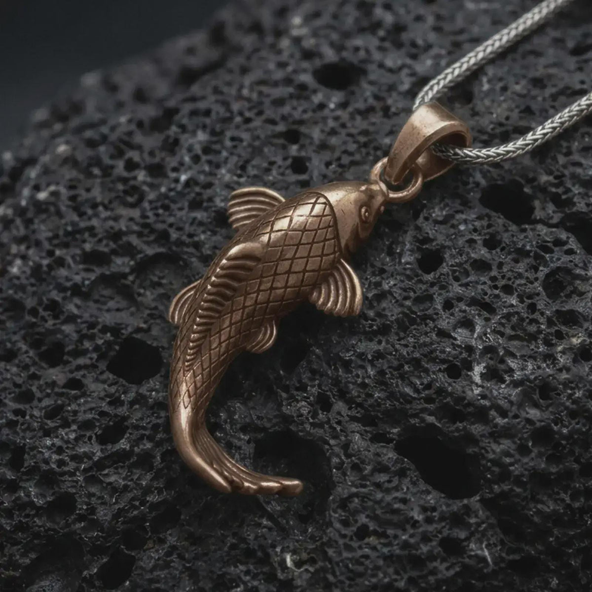 Bronze Japanese koi fish pendant on black volcanic rock, featuring engraved diamond scales, flowing fins, a curved body, and detailed fish tail.