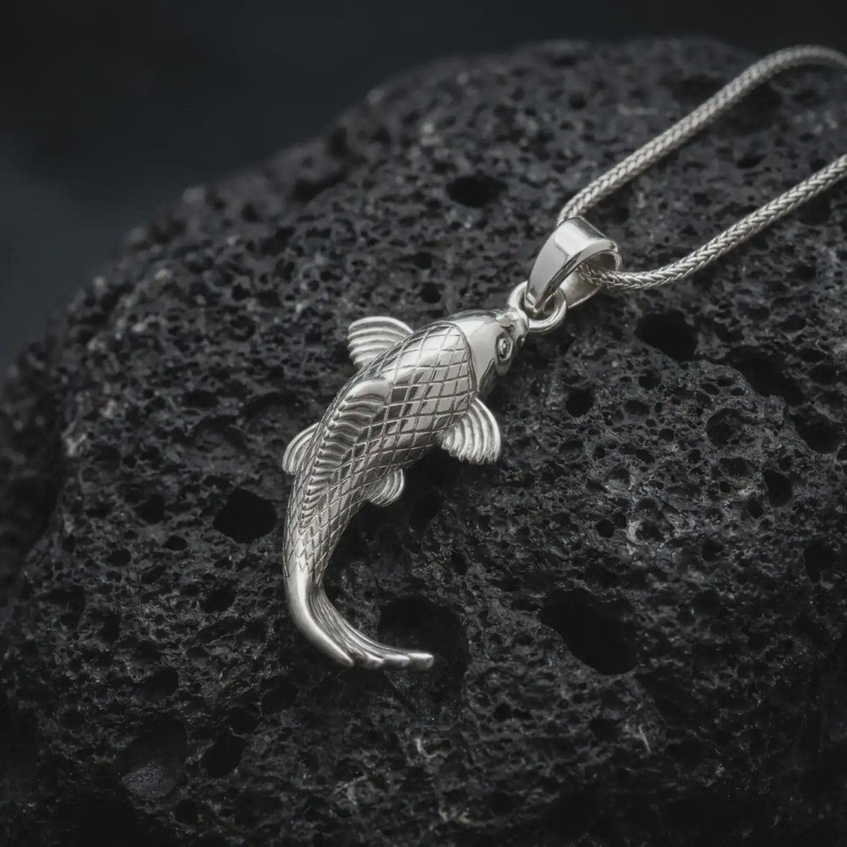 Polished sterling silver koi necklace displayed on dark volcanic stone, highlighting the sculpted carp body, engraved scale pattern, flowing fins, and graceful swimming shape.