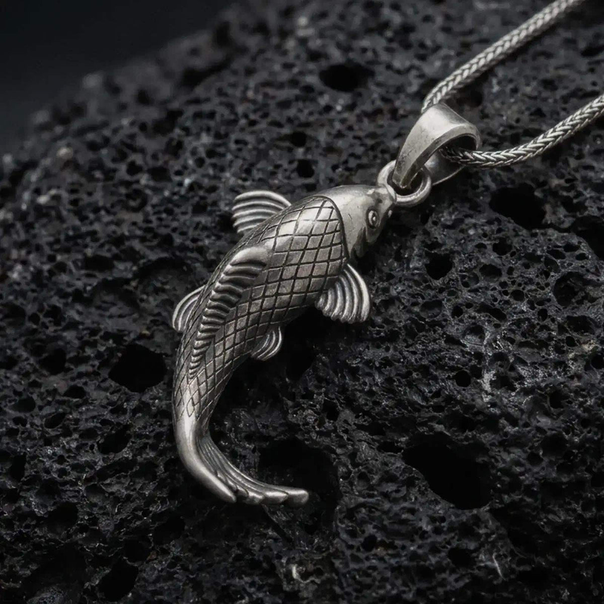 Oxidized sterling silver Japanese koi fish pendant on black volcanic rock, featuring engraved diamond scales, flowing fins, a curved body, and detailed fish tail.