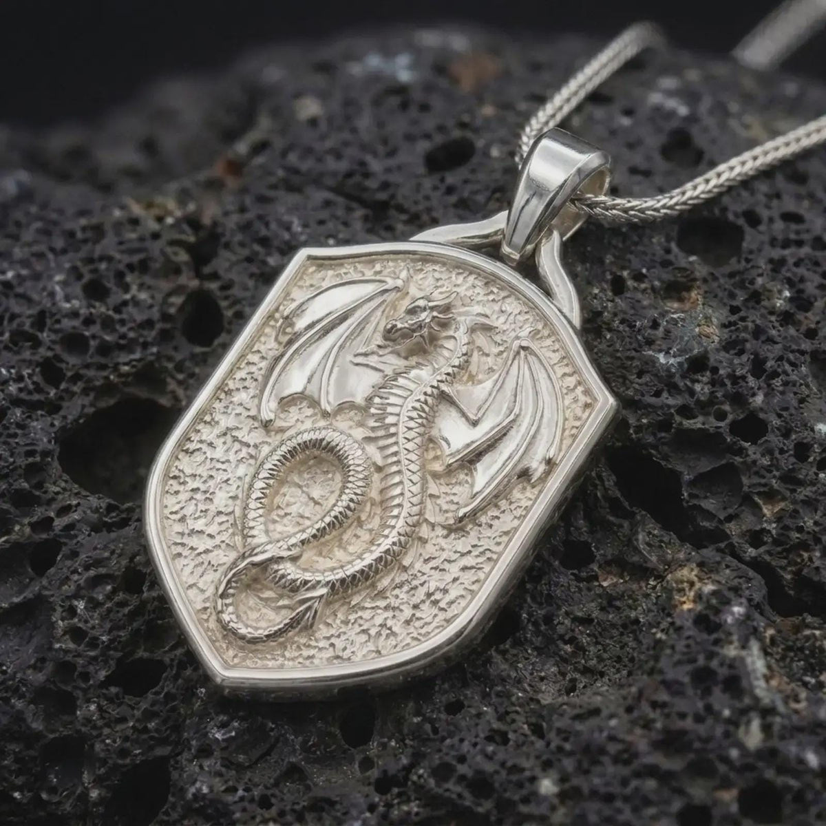 Polished sterling silver dragon shield necklace displayed on dark volcanic stone, highlighting the sculpted wings, scaled coiled body, fierce dragon head, and heraldic shield frame.