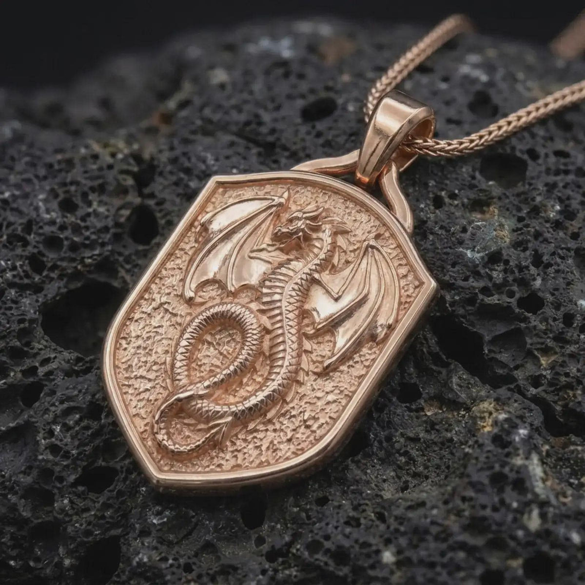 Rose gold plated dragon shield necklace displayed on textured volcanic stone, showcasing the winged dragon relief, serpentine body, detailed scales, and gothic fantasy styling.