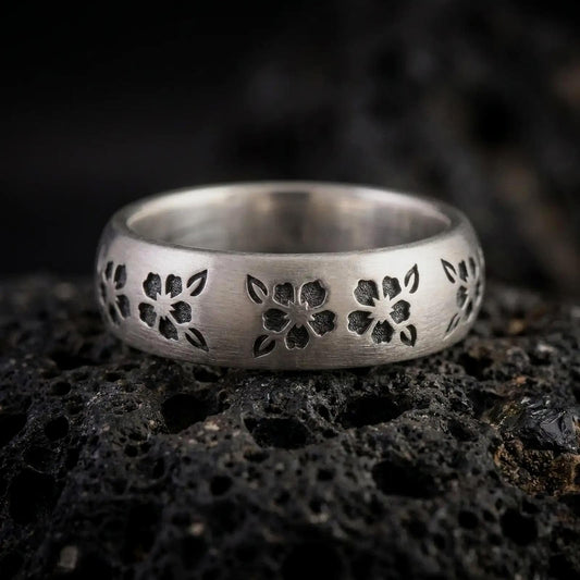 Oxidized sterling silver sakura ring on black volcanic rock, featuring recessed five-petal cherry blossoms, small leaves, dark textured details, and a wide Japanese floral band.