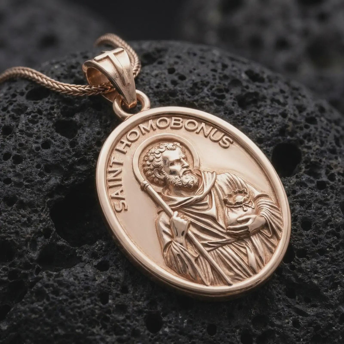 Rose gold plated Saint Homobonus necklace displayed on textured volcanic stone, showcasing the haloed saint, staff, devotional imagery, and oval religious medal.