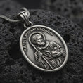 Load image into Gallery viewer, Oxidized sterling silver Saint Homobonus medal necklace on black volcanic rock, featuring the haloed Catholic saint holding a staff with SAINT HOMOBONUS lettering.
