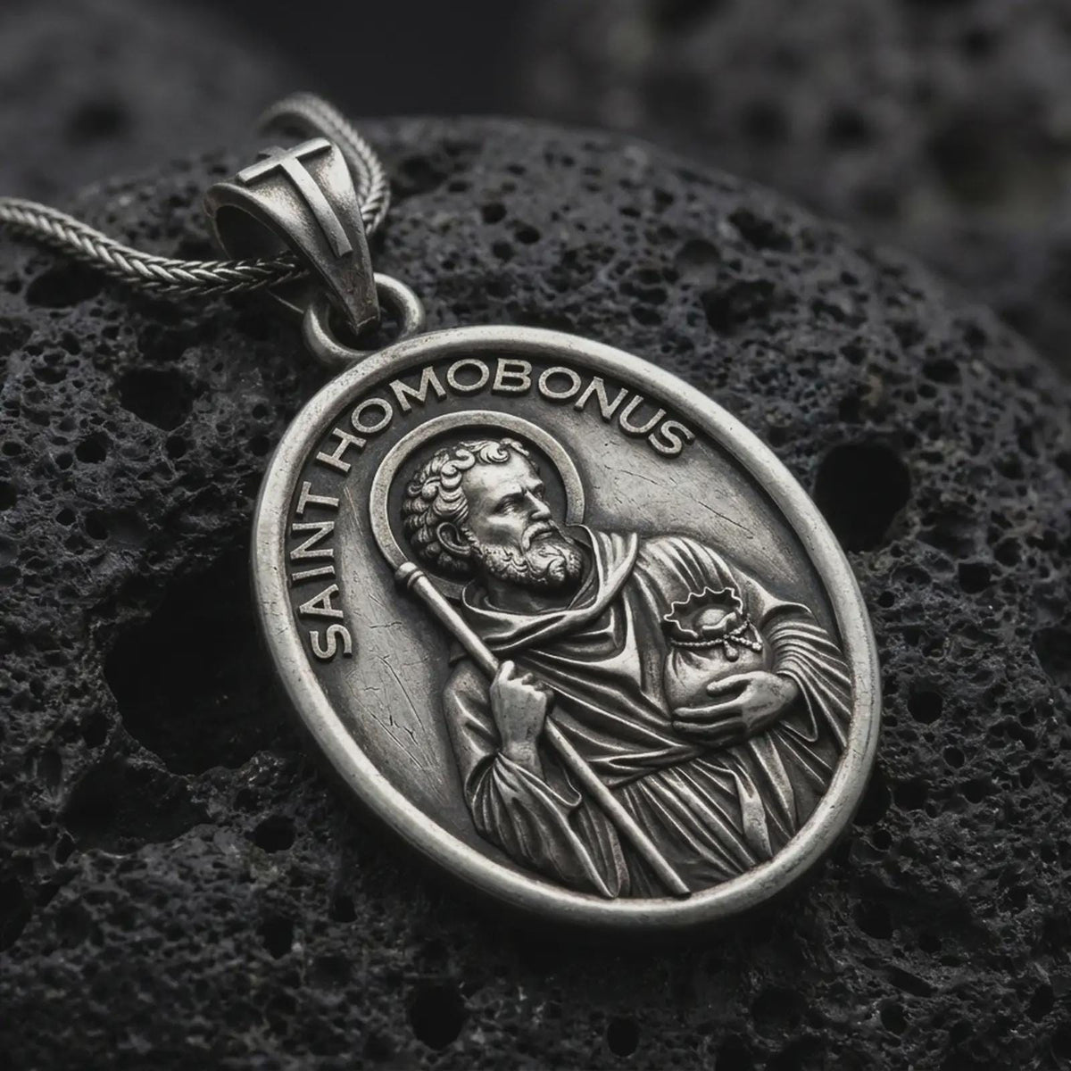 Oxidized sterling silver Saint Homobonus medal necklace on black volcanic rock, featuring the haloed Catholic saint holding a staff with SAINT HOMOBONUS lettering.