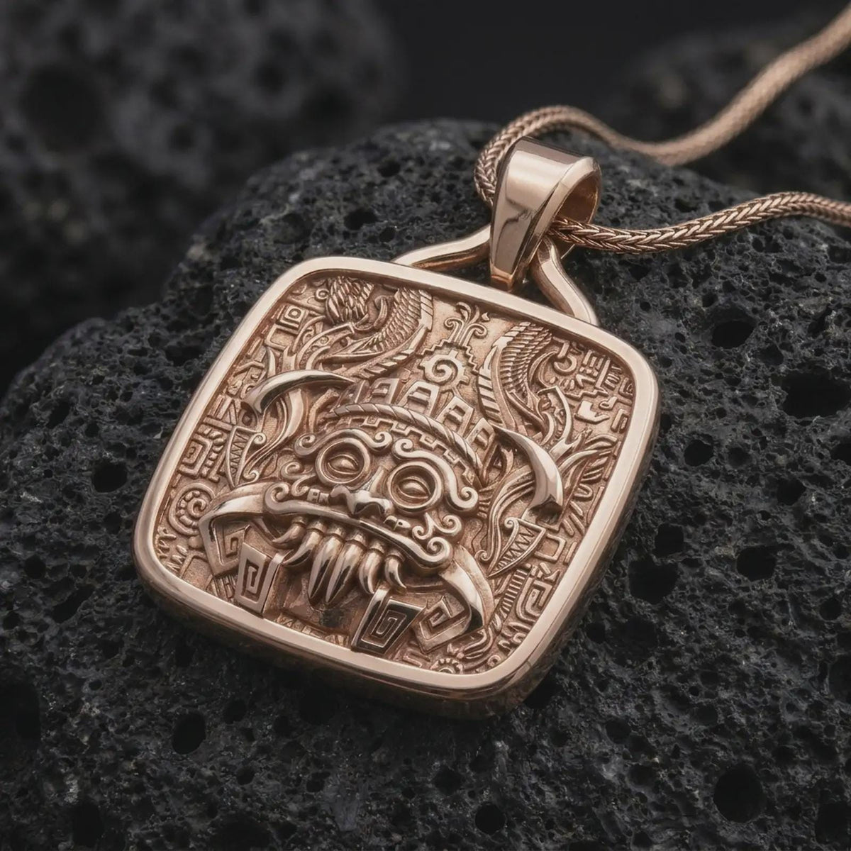 Rose gold plated Tlaloc necklace displayed on textured volcanic stone, showcasing the fierce rain god mask, geometric Aztec patterns, ceremonial details, and square pendant frame.