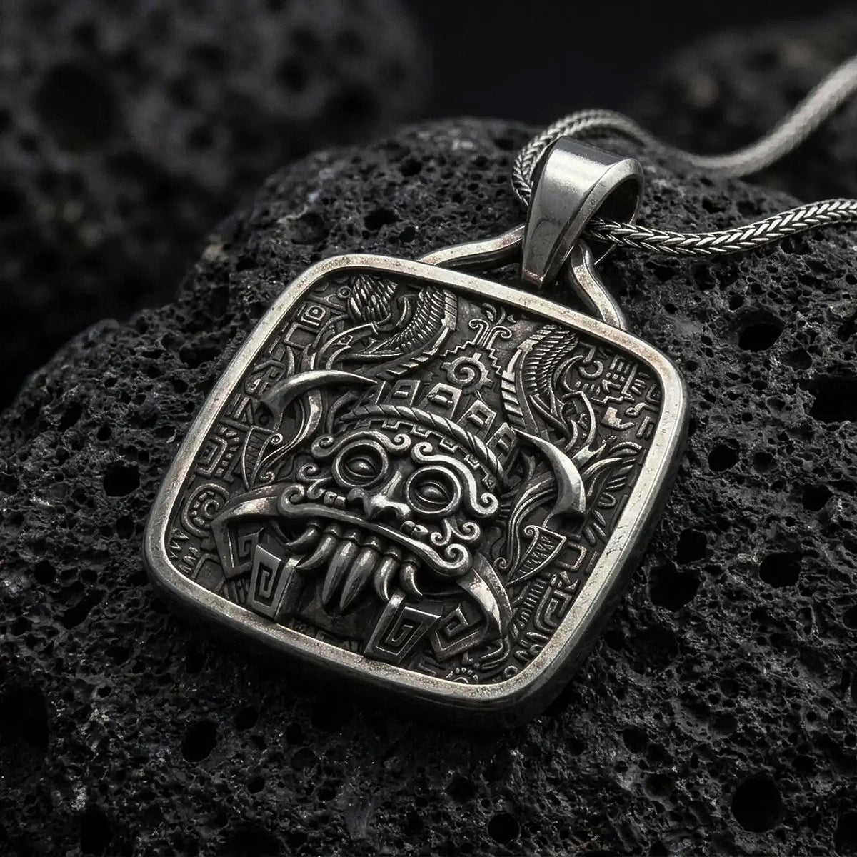Oxidized sterling silver Tlaloc pendant on black volcanic rock, featuring ringed eyes, prominent fangs, curled facial details, and intricate Aztec geometric patterns.