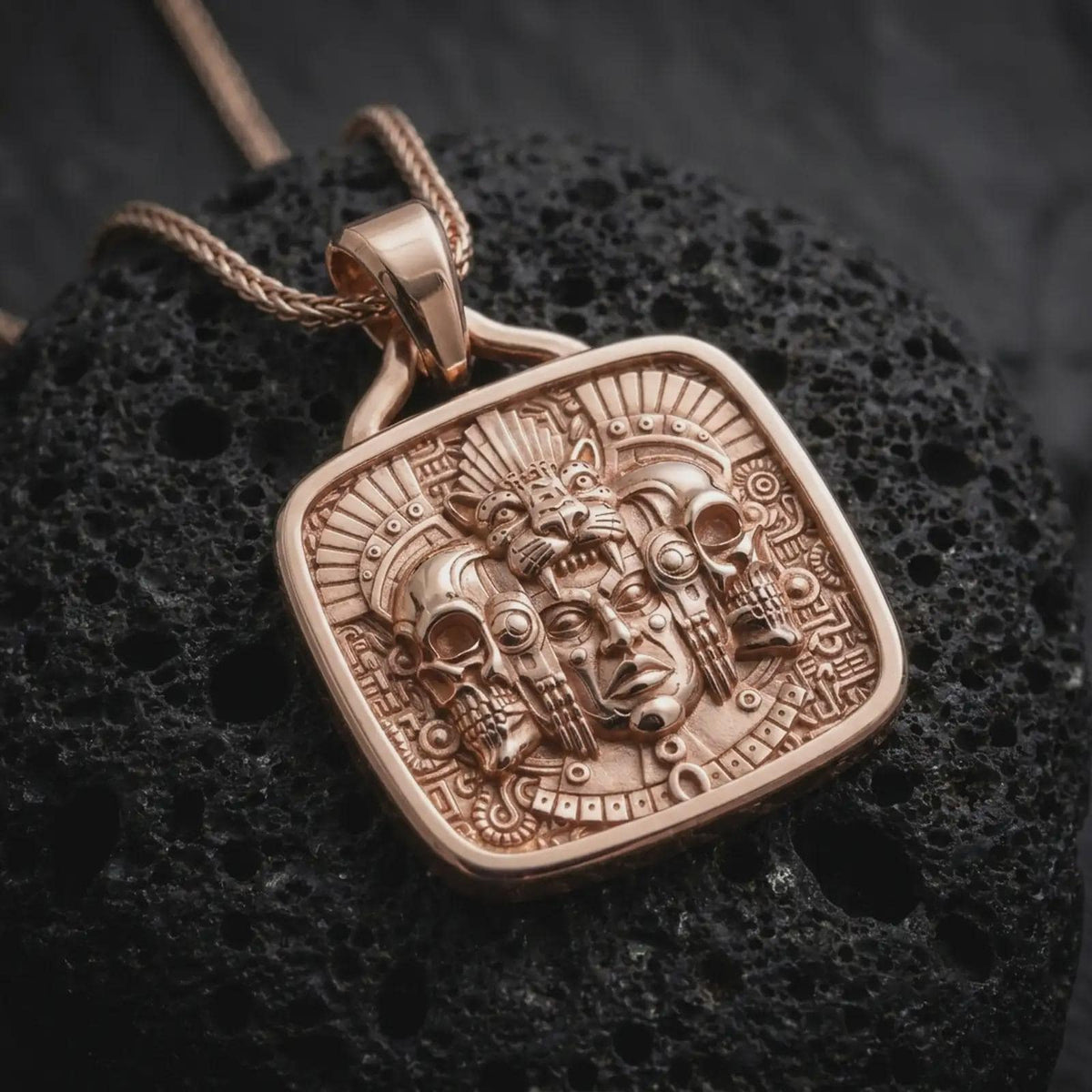 Rose gold plated Mexica pendant displayed on textured volcanic stone, showcasing the detailed jaguar headdress, human face, skull ornaments, feathers, and bold Aztec-inspired relief.