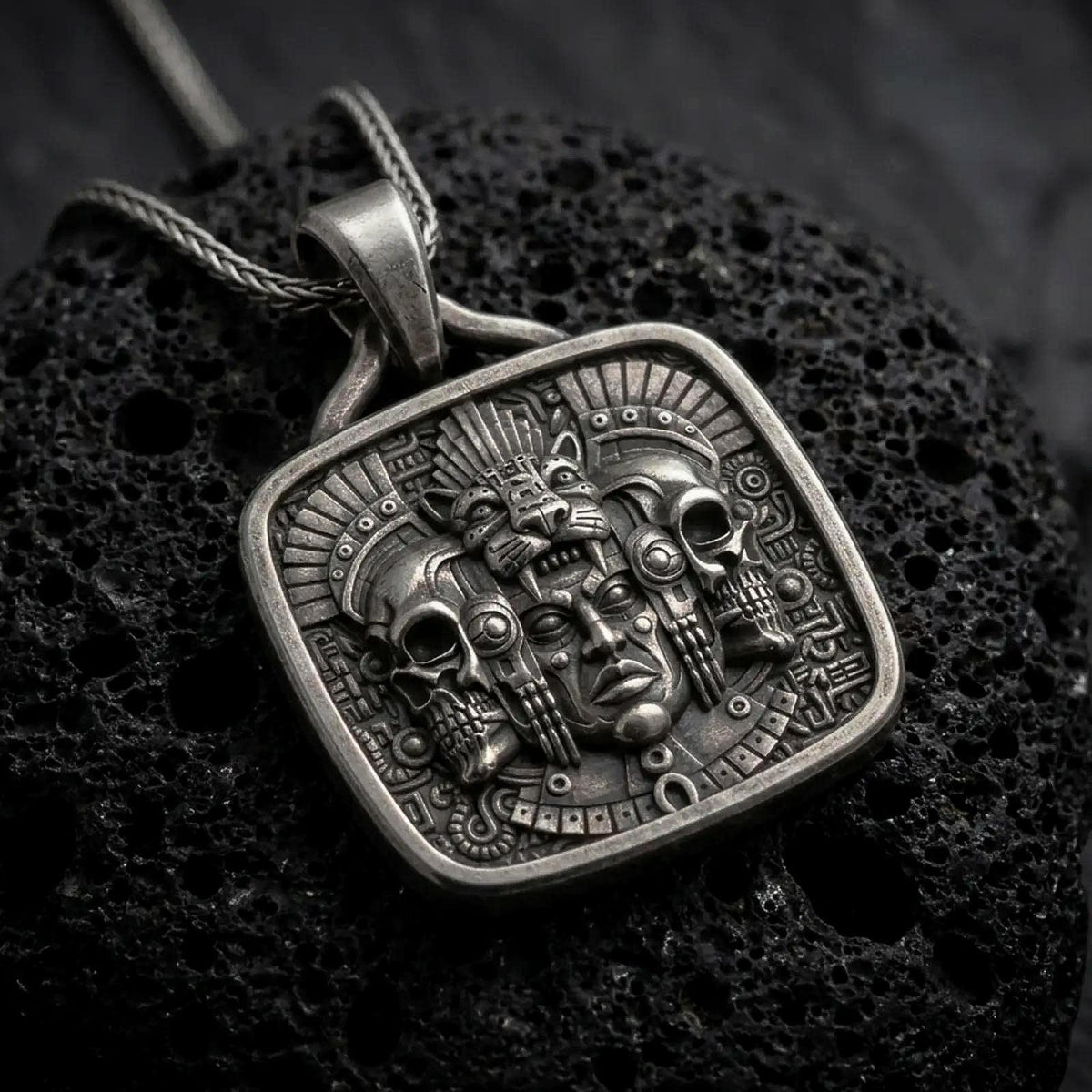 Oxidized sterling silver Aztec jaguar pendant on black volcanic rock, featuring a ceremonial human face, feline headdress, flanking skulls, feathers, and intricate Mesoamerican relief details.
