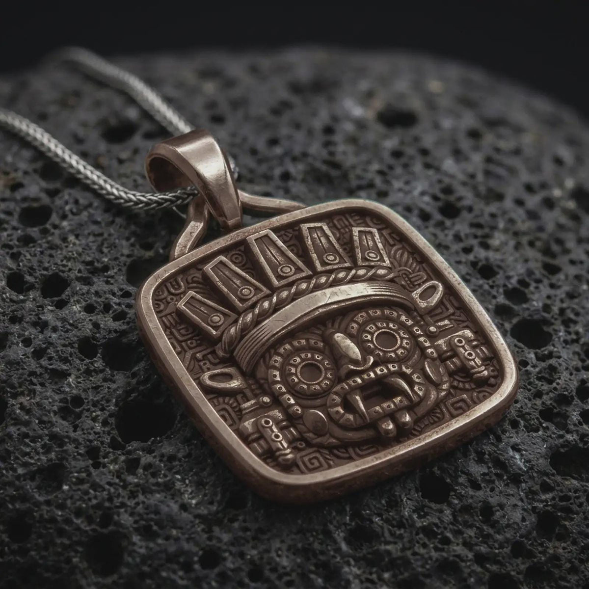 Bronze Tlaloc pendant on black volcanic rock, featuring iconic goggle eyes, fanged mouth, ceremonial headdress, and detailed Aztec geometric patterns.