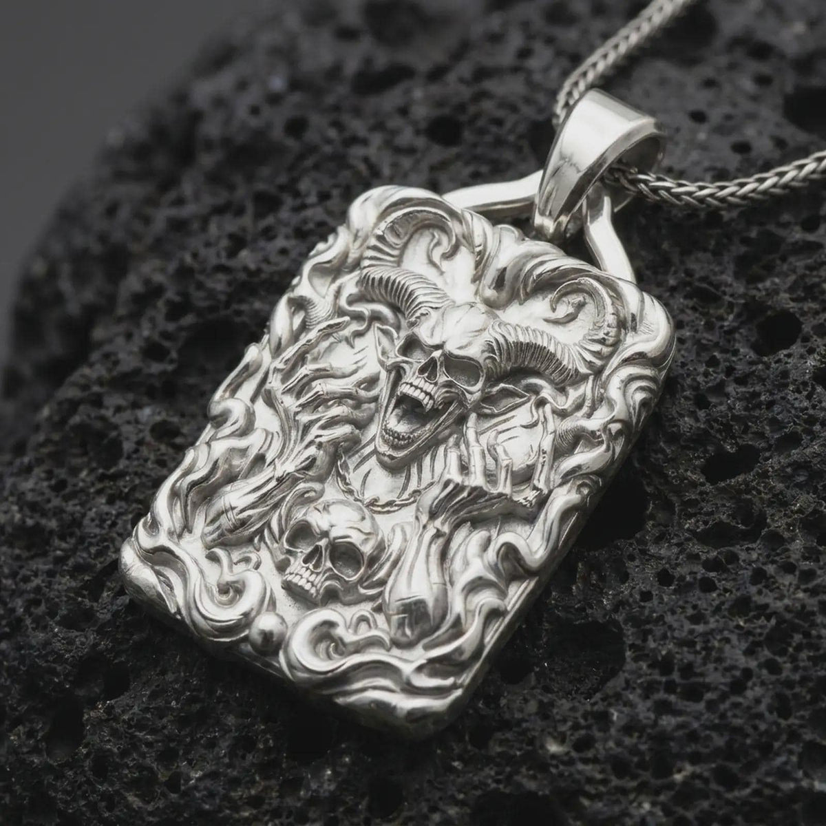Polished sterling silver demon skull necklace displayed on dark volcanic stone, highlighting the horned skull, skeletal hands, flame details, and dramatic dark fantasy design.