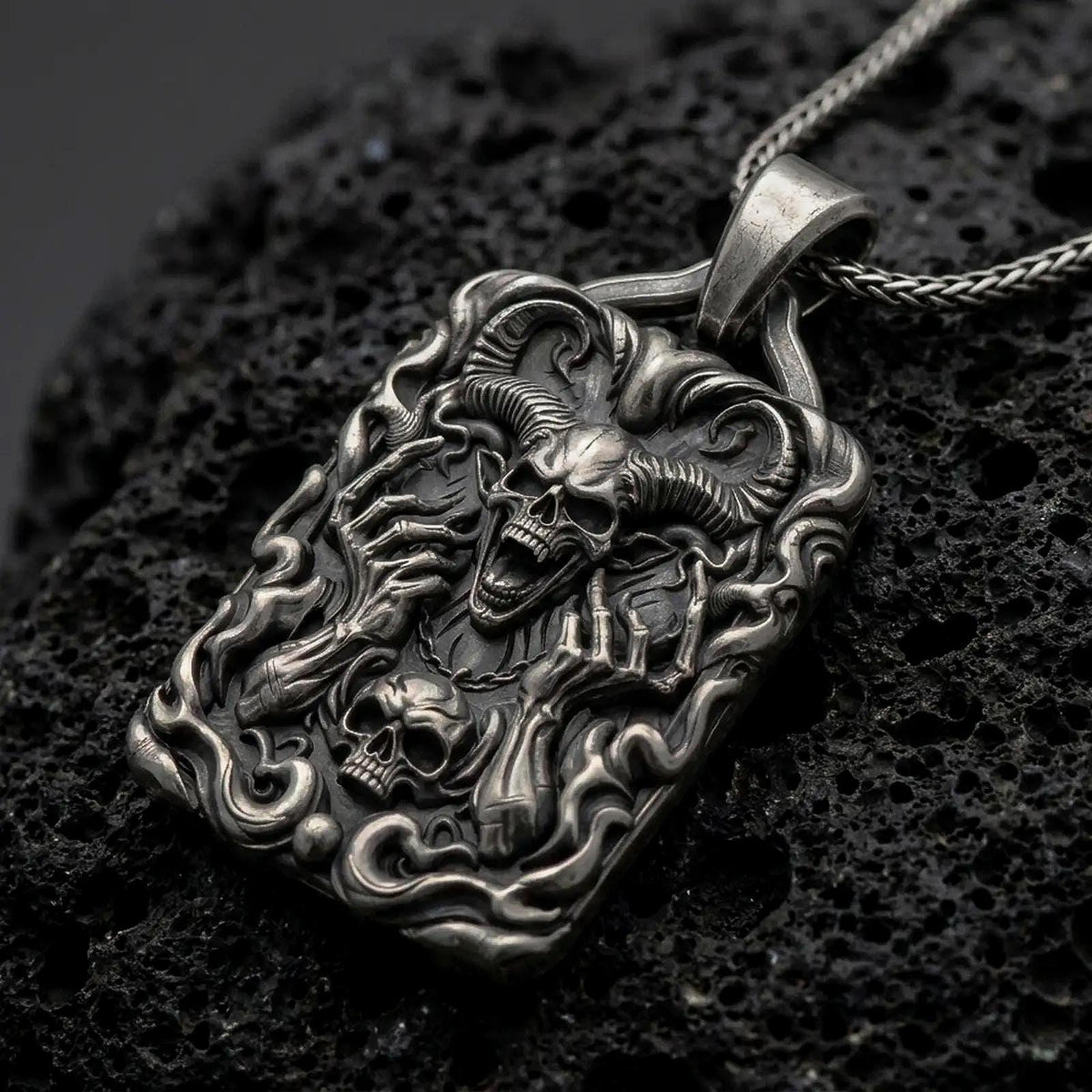Oxidized sterling silver horned demon skull pendant on black volcanic rock, featuring large curled ram horns, skeletal hands, flames, and a smaller skull in detailed gothic relief.