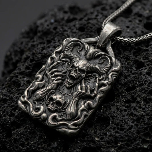 Oxidized sterling silver horned demon skull pendant on black volcanic rock, featuring large curled ram horns, skeletal hands, flames, and a smaller skull in detailed gothic relief.