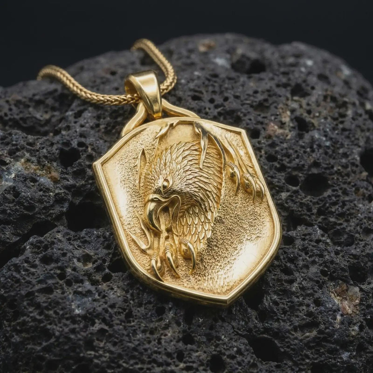Gold plated eagle shield pendant resting on black volcanic rock, featuring a powerful eagle head, detailed plumage, sharp claws, and bold heraldic medallion design.