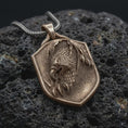 Load image into Gallery viewer, Bronze eagle shield pendant on black volcanic rock, featuring a detailed eagle head, layered feathers, sharp talons, and textured heraldic shield.
