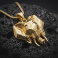 Load image into Gallery viewer, Gold plated elephant head pendant resting on black volcanic rock, featuring detailed African elephant styling, large ears, prominent tusks, and a bold dimensional animal design.
