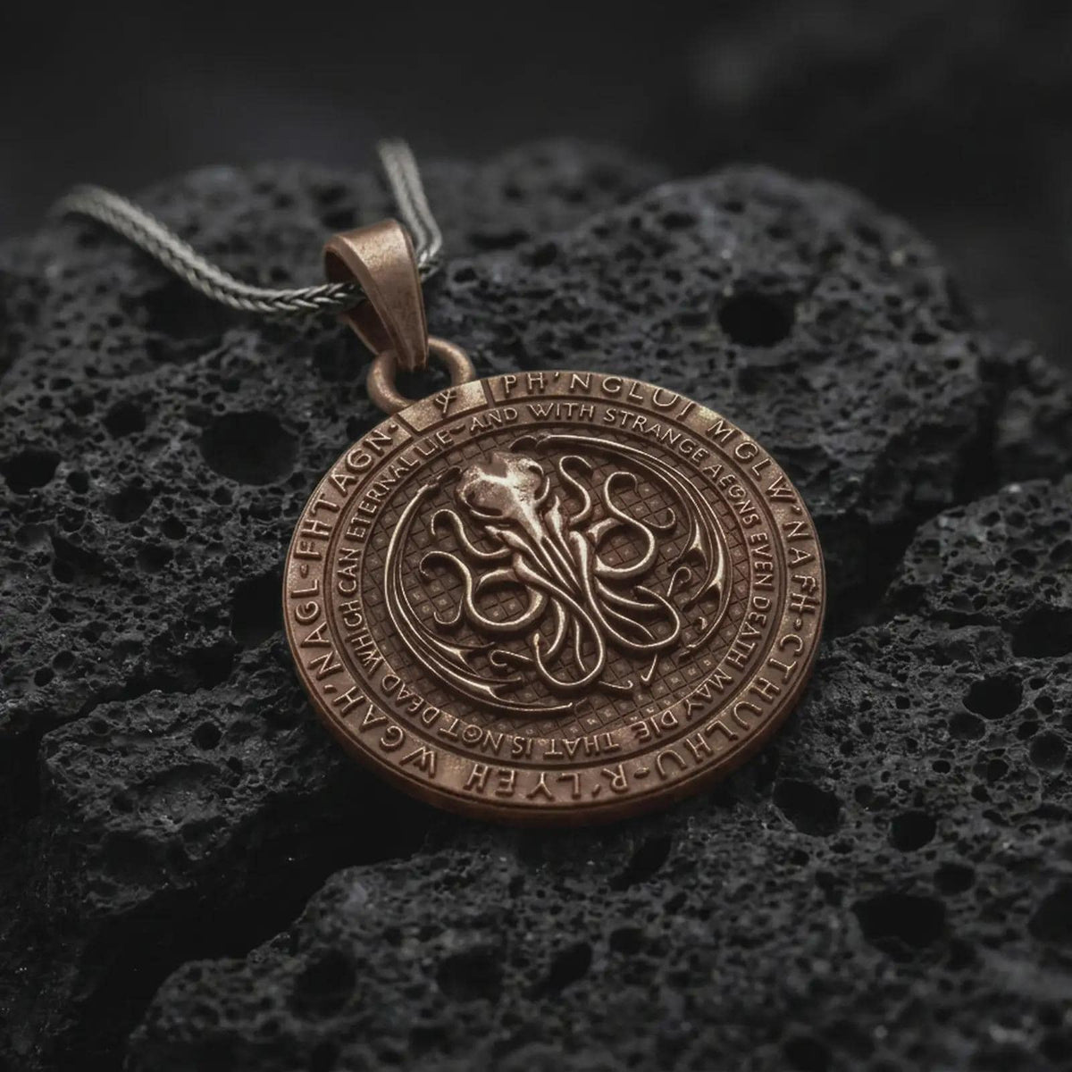 Bronze Cthulhu pendant on black volcanic rock, featuring a raised cephalopod sigil in the center, engraved outer ring lettering, and a dark Lovecraftian medallion design.