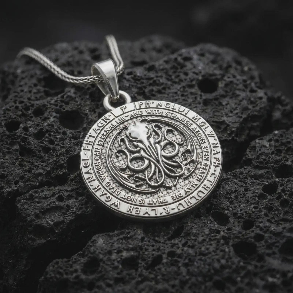 Polished sterling silver Lovecraft necklace displayed on textured volcanic stone, highlighting the circular pendant shape, tentacled Cthulhu motif, and engraved mythos-inspired border text.