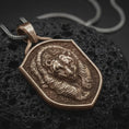 Load image into Gallery viewer, Bronze grizzly bear shield pendant on black volcanic rock, featuring a sculpted bear head, layered fur relief, textured background, and raised shield border.
