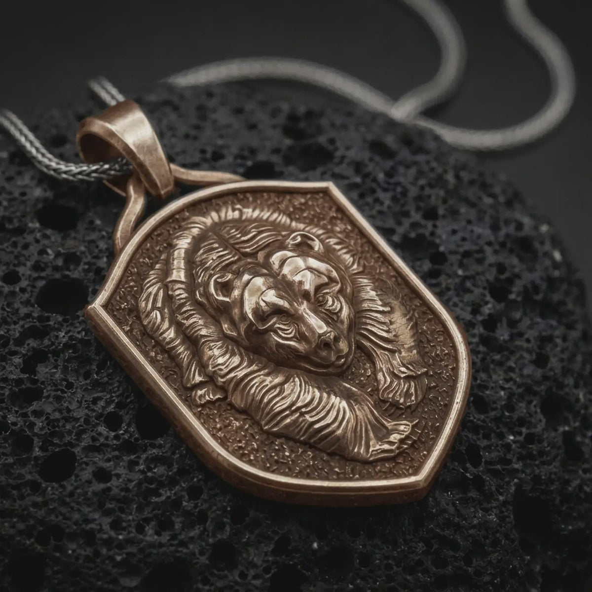 Bronze grizzly bear shield pendant on black volcanic rock, featuring a sculpted bear head, layered fur relief, textured background, and raised shield border.