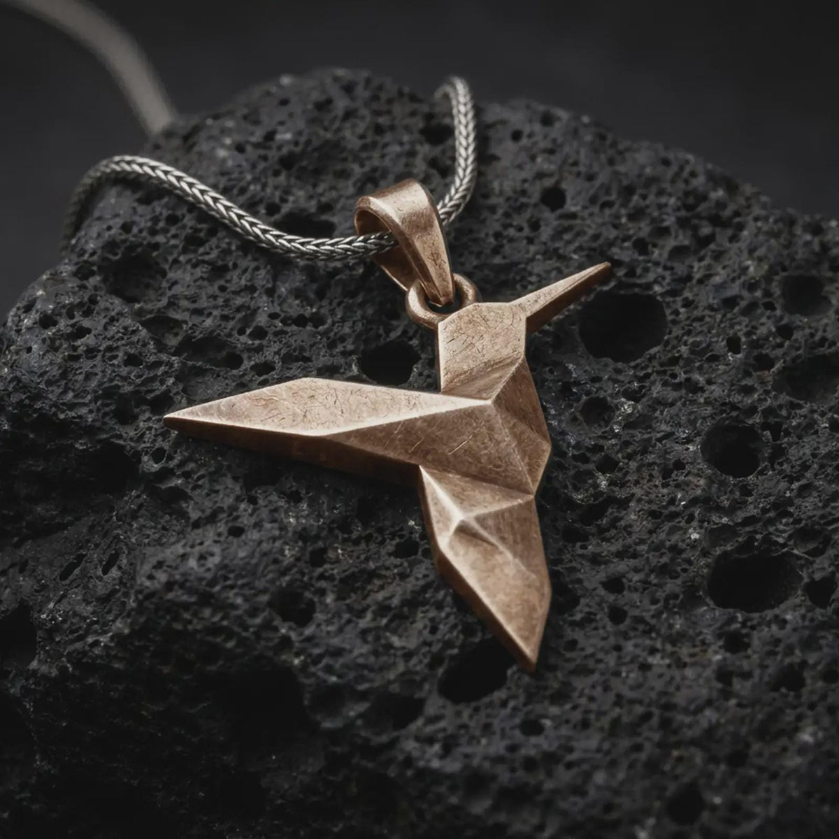 Bronze origami hummingbird pendant on black volcanic rock, featuring a geometric folded bird design with a long pointed beak and angular wings.