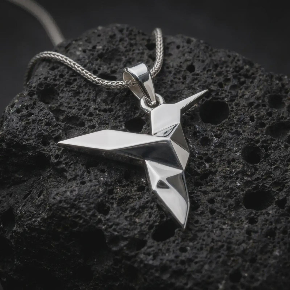 Polished sterling silver hummingbird necklace displayed on dark volcanic stone, highlighting the minimalist origami bird form, sharp edges, and modern sculptural silhouette.