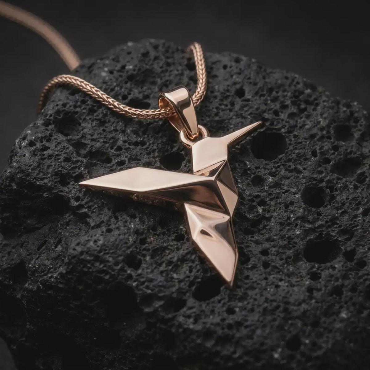 Rose gold plated hummingbird necklace on textured volcanic stone, featuring a folded origami style bird pendant with angular body lines and a minimalist elegant finish.