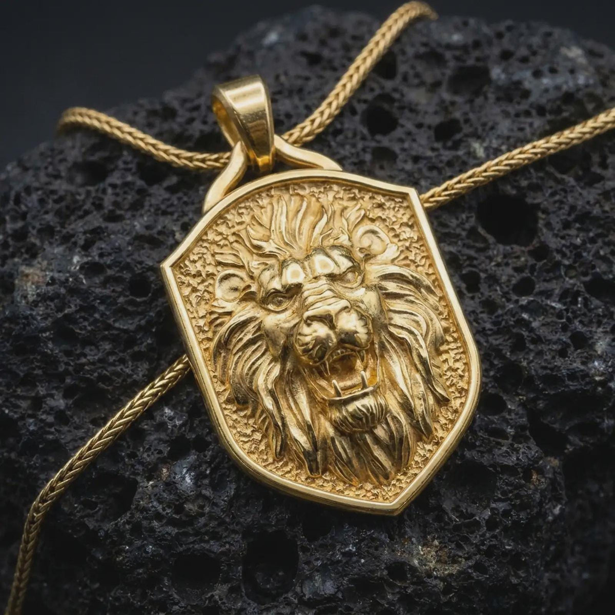 Gold plated roaring lion pendant resting on black volcanic rock, featuring an aggressive lion portrait, sharp canine teeth, flowing mane, and bold medieval-inspired shield design.