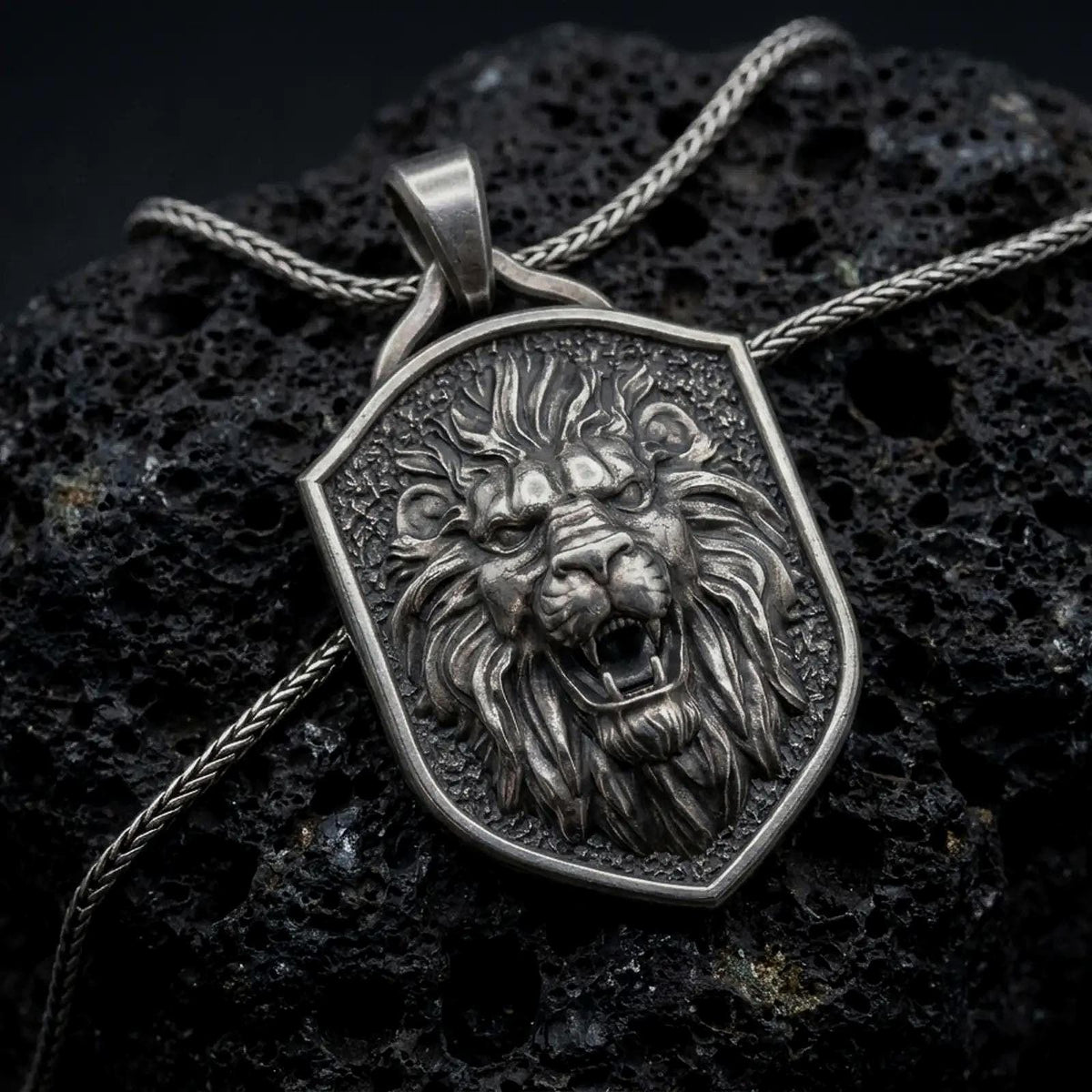 Oxidized sterling silver roaring lion shield pendant on black volcanic rock, featuring a fierce lion head, open jaws, sharp fangs, flowing mane, and textured heraldic shield background.