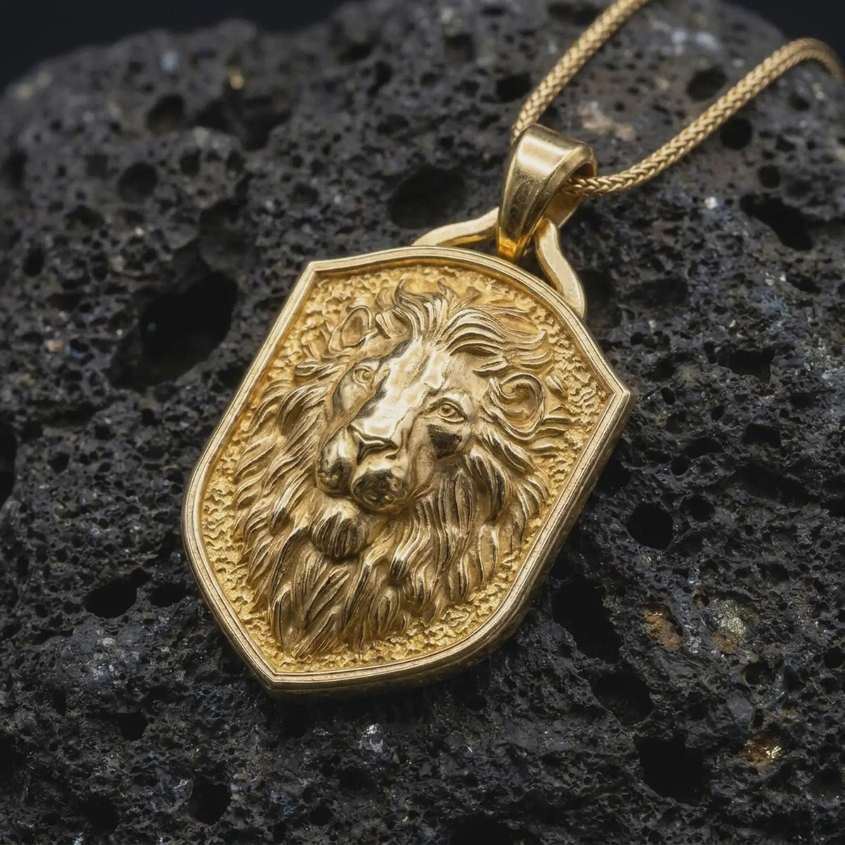 Gold plated regal lion pendant resting on black volcanic rock, featuring a majestic lion head, layered mane, textured background, and bold heraldic shield silhouette.