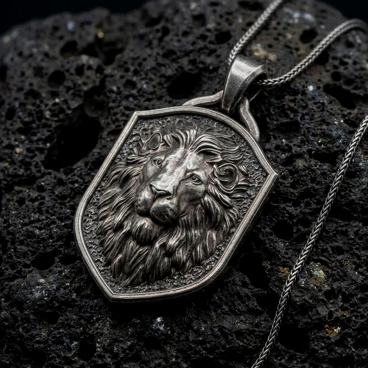 Oxidized sterling silver regal lion shield pendant on black volcanic rock, featuring a majestic lion portrait, calm expression, flowing mane, and textured heraldic shield background.