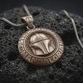 Load image into Gallery viewer, Bronze Mandalorian helmet pendant on black volcanic rock, featuring a raised bounty hunter helmet, circular Star Wars lettering, and engraved insignia bail.
