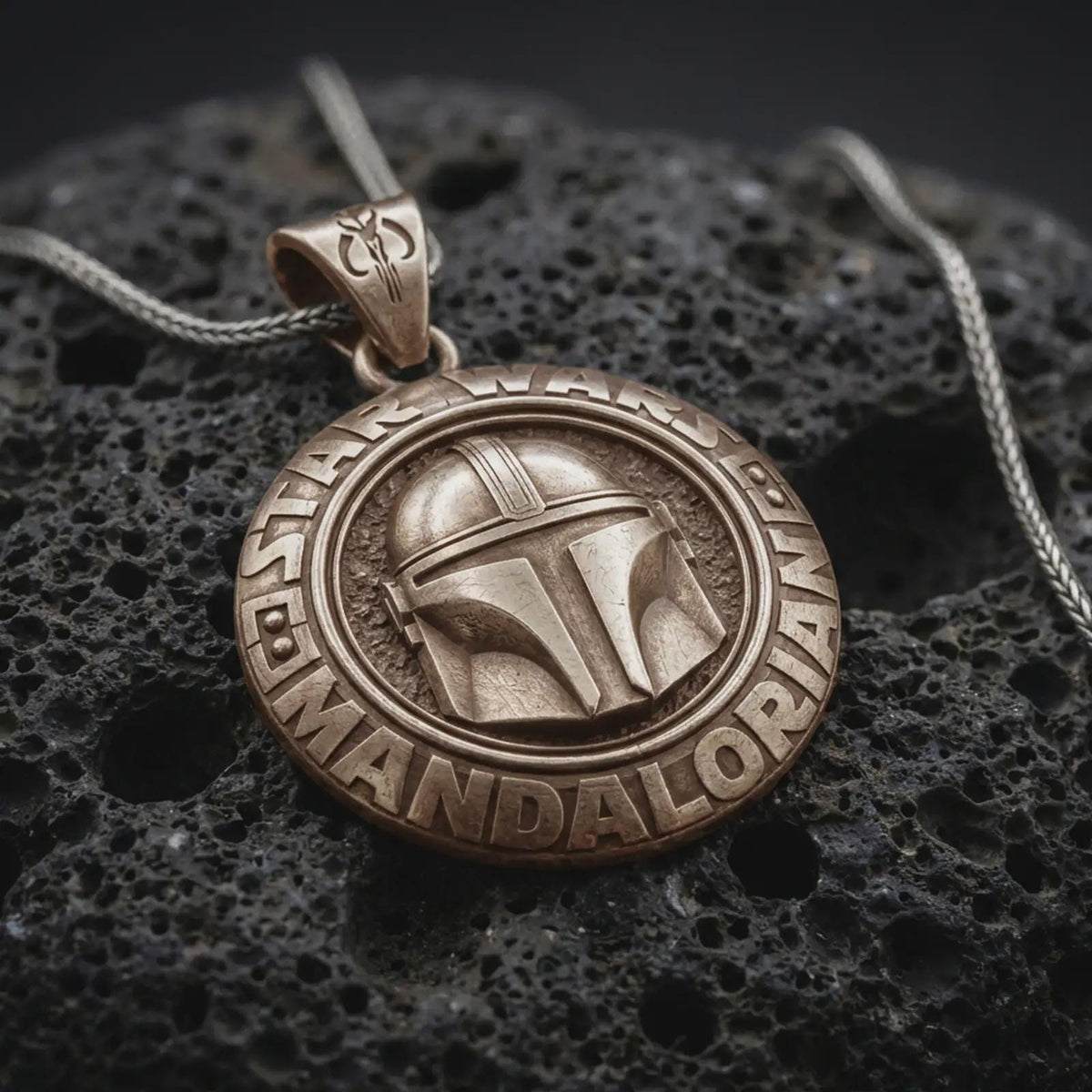 Bronze Mandalorian helmet pendant on black volcanic rock, featuring a raised bounty hunter helmet, circular Star Wars lettering, and engraved insignia bail.