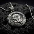 Load image into Gallery viewer, Oxidized sterling silver Mandalorian helmet pendant on black volcanic rock, featuring a raised bounty hunter helmet, circular Star Wars lettering, and engraved insignia bail.
