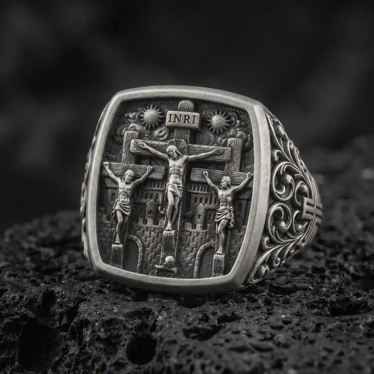 Oxidized sterling silver Crucifixion ring on black volcanic rock, featuring Jesus Christ on the center cross between the two thieves, INRI inscription, and detailed Calvary scene.