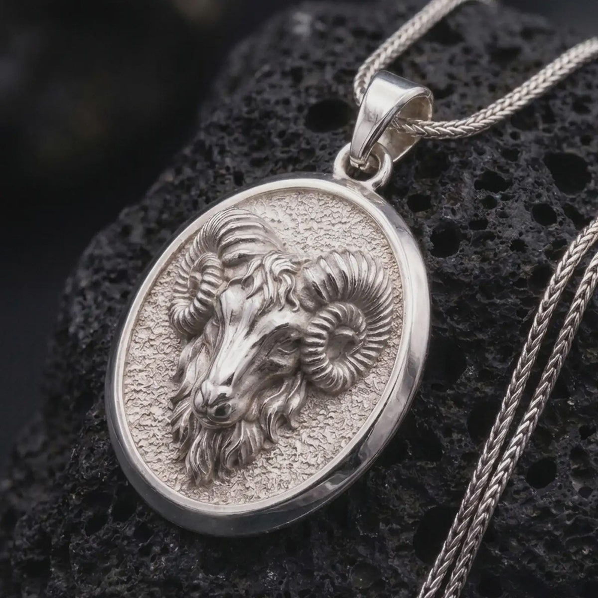Polished sterling silver ram head necklace displayed on dark volcanic stone, highlighting the dimensional face, symmetrical curled horns, detailed fur, and oval zodiac pendant.