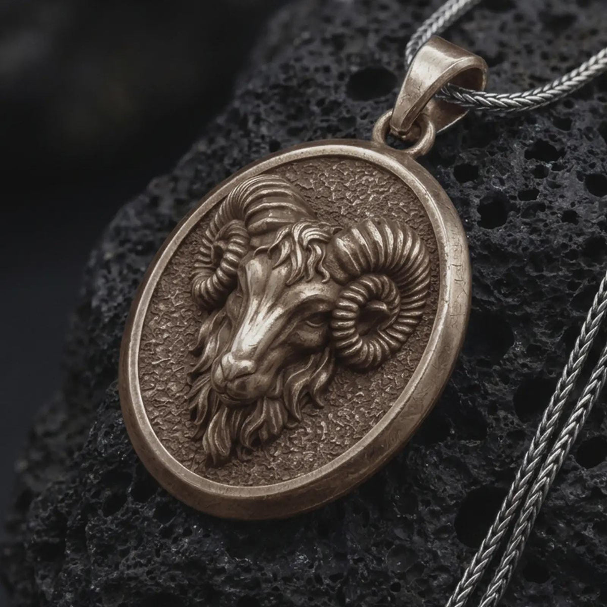 Bronze Aries ram pendant on black volcanic rock, featuring a sculpted ram head, large curled horns, textured background, and oval medallion frame.