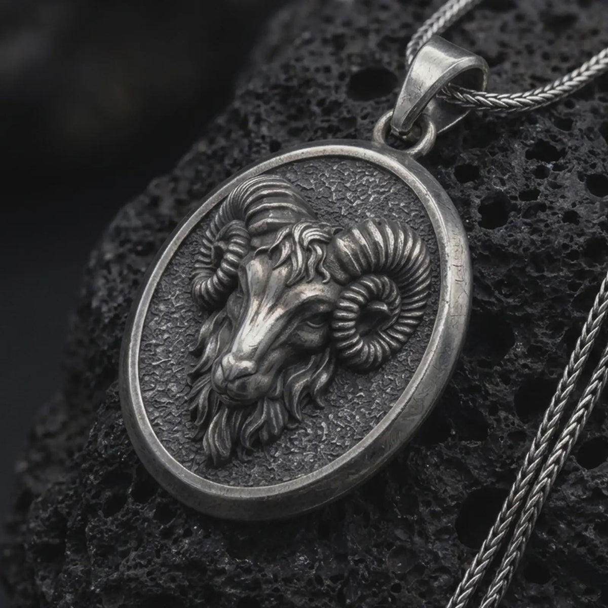 Oxidized sterling silver Aries ram pendant on black volcanic rock, featuring a sculpted ram head, large curled horns, textured background, and oval medallion frame.