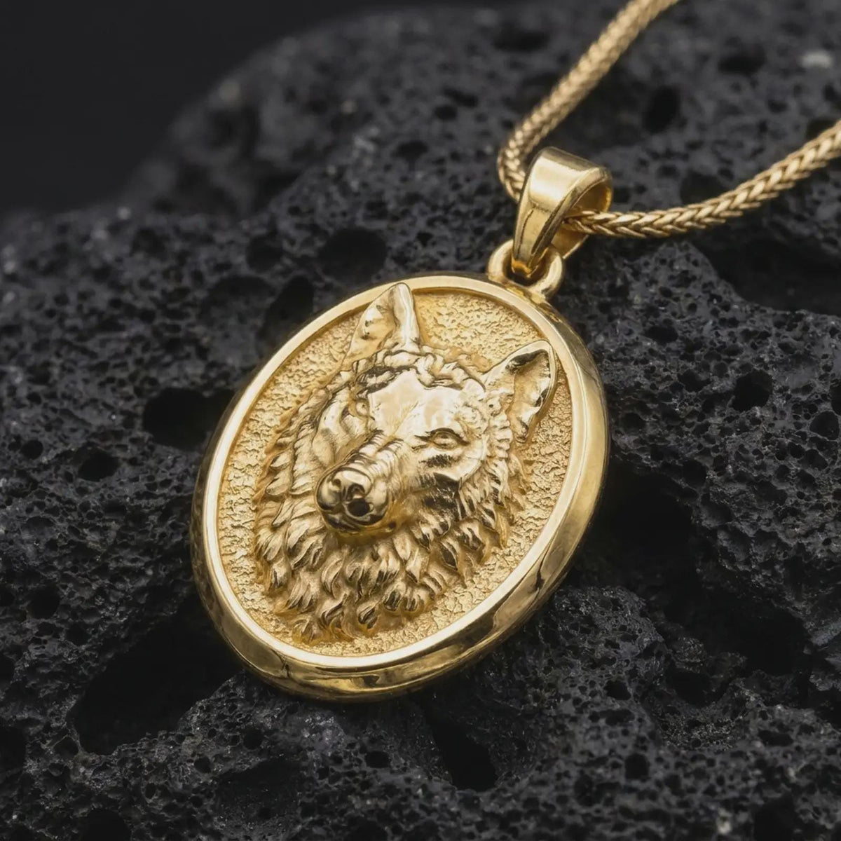 Gold plated wolf head pendant resting on black volcanic rock, featuring a detailed wolf face, textured fur, pointed ears, and bold oval wildlife necklace design.