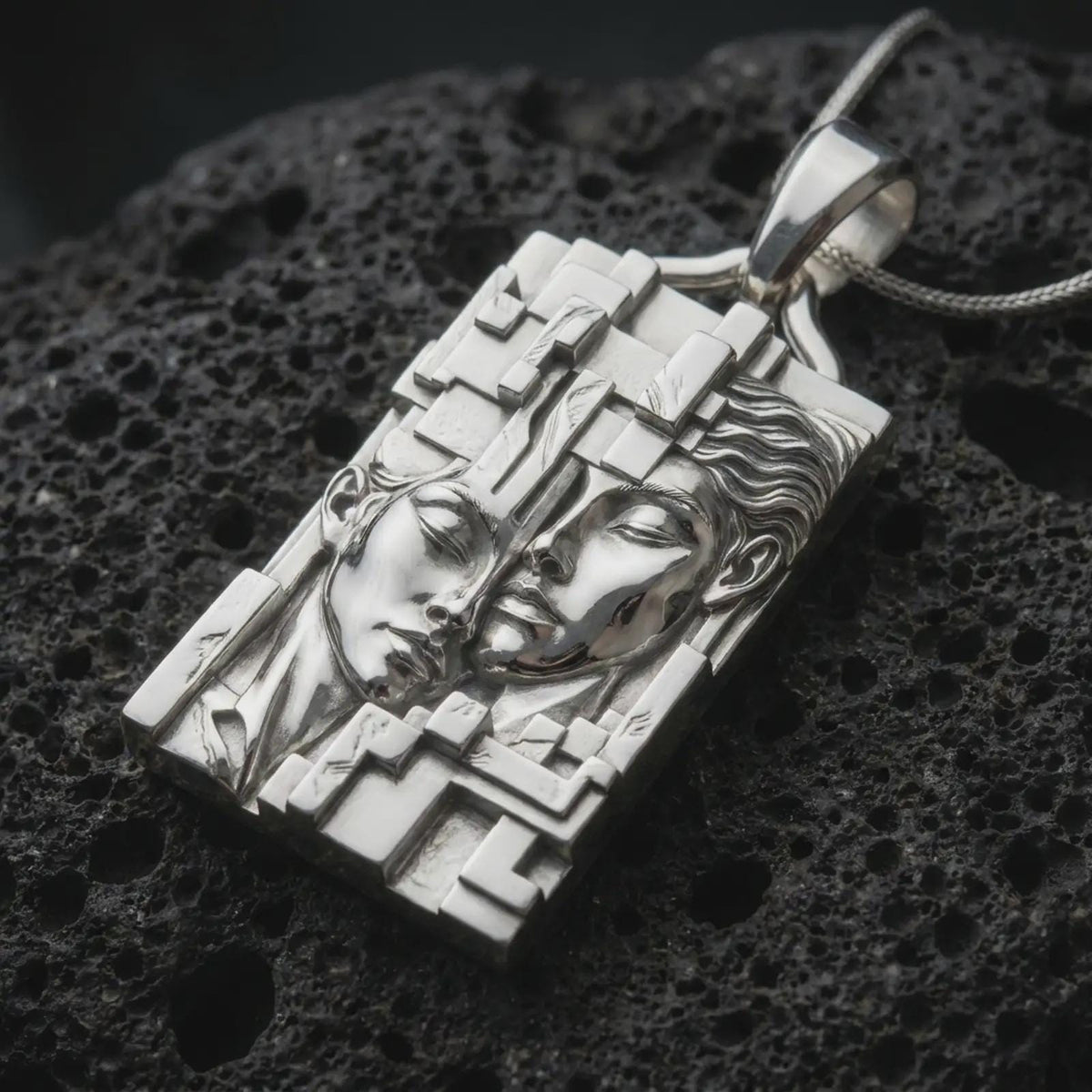 Polished sterling silver Gemini zodiac necklace displayed on dark volcanic stone, highlighting the twin face relief, flowing hair details, angular architectural borders, and dimensional silver design.